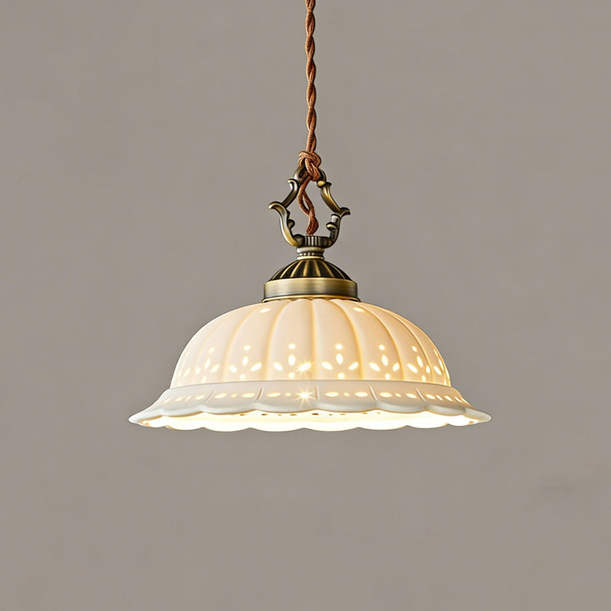 Lit pierced ceramic dome of this Clérachic pendant glowing cream on a grey wall, with an ornate antique brass cap above.