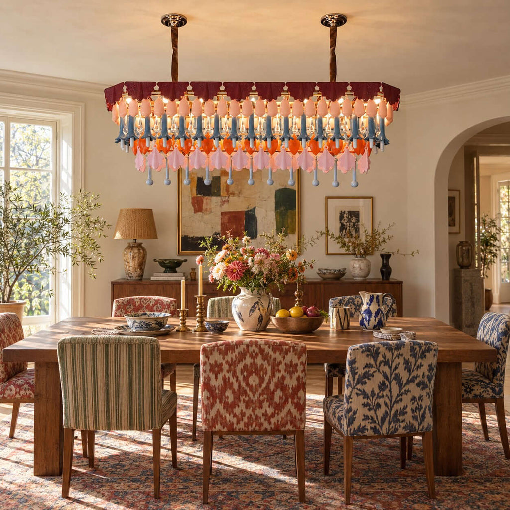 A rectangular Clérachic chandelier hangs over a rustic dining table with patterned chairs, with a lamp and flowers on a sideboard.