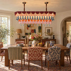 A rectangular Clérachic chandelier hangs over a rustic dining table with patterned chairs, with a lamp and flowers on a sideboard.