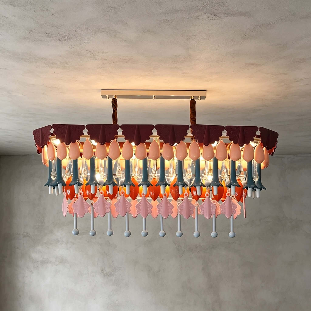 A rectangular Clérachic chandelier on a straight ceiling bar, with pink, orange and teal drops lit warmly against grey plaster.