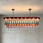 A long Clérachic chandelier hung from two chains and canopies, with rows of teal, orange and pink drops lit on a grey wall.