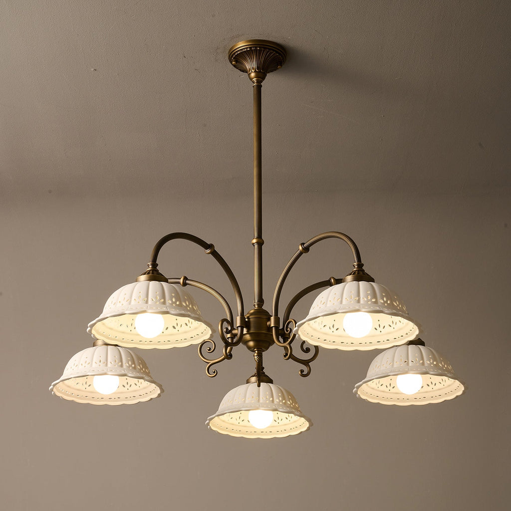 Underside view of this lit Clérachic chandelier, five white pierced ceramic shades on antique brass arms against a taupe wall.