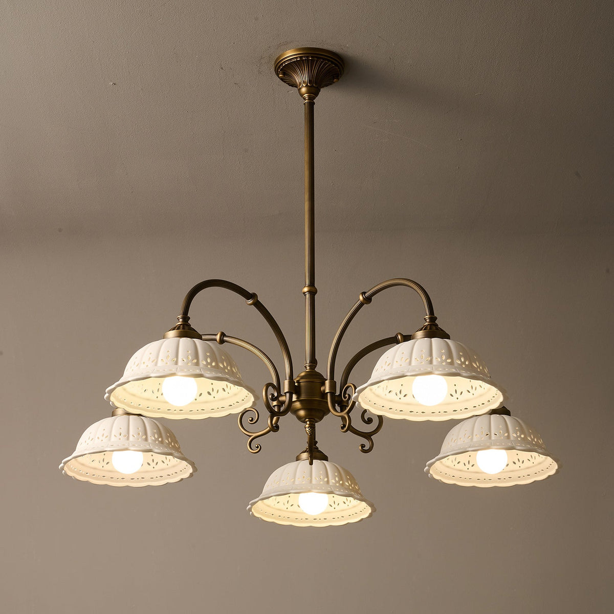 Underside view of this lit Clérachic chandelier, five white pierced ceramic shades on antique brass arms against a taupe wall.