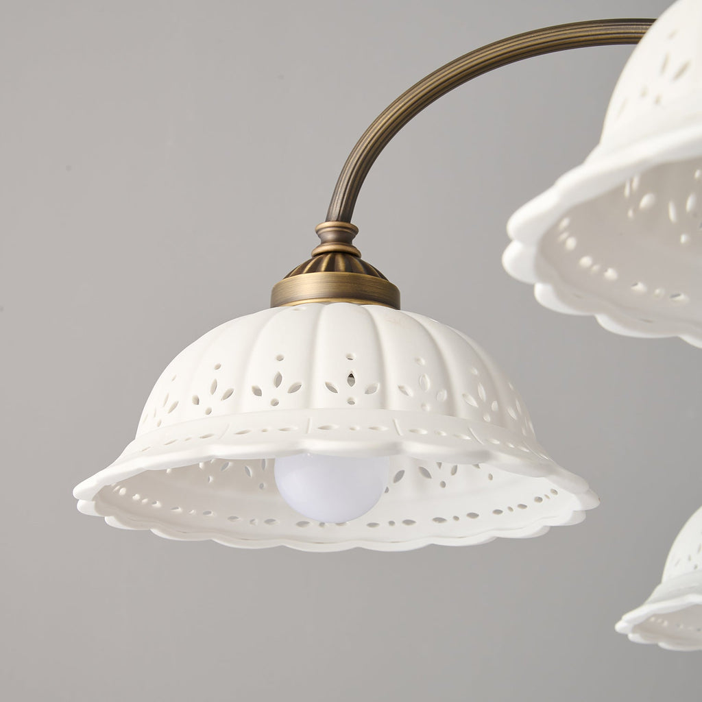 Close view of one white ceramic shade with a pierced leaf pattern and scalloped edge on this Clérachic chandelier, a bulb glowing inside.