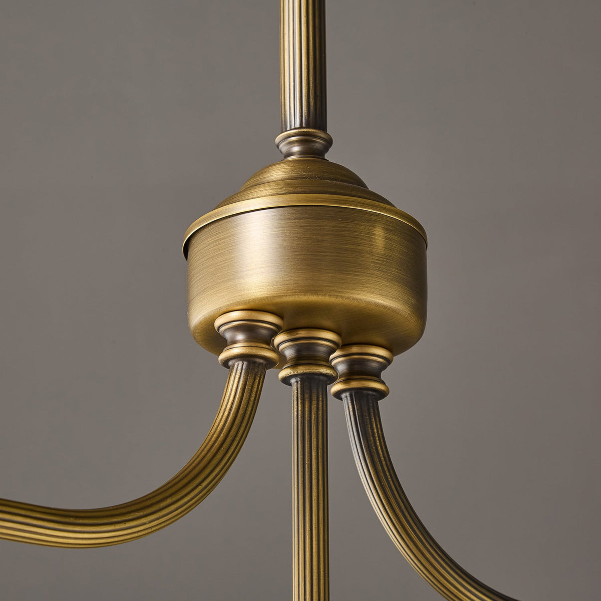 Close view of the round antique brass hub with fluted arms on this Clérachic chandelier, against a grey wall.