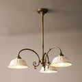 Three-light round Clérachic chandelier on a brass rod, its white pierced shades lit and curved arms spreading below a taupe ceiling.