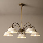 Lit Clérachic five-light chandelier on a long brass rod, hung against a taupe ceiling, with white ceramic shades on curved arms.
