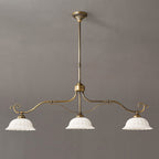 Linear Clérachic chandelier with three white ceramic shades on a long, gently curved brass bar, hung from a rod against a grey wall.