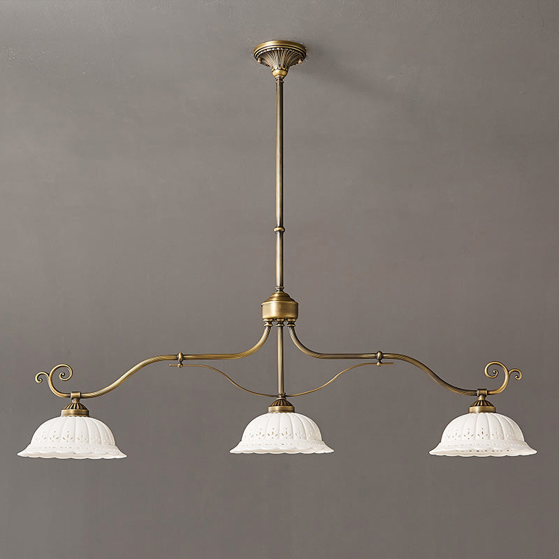 Linear Clérachic chandelier with three white ceramic shades on a long, gently curved brass bar, hung from a rod against a grey wall.