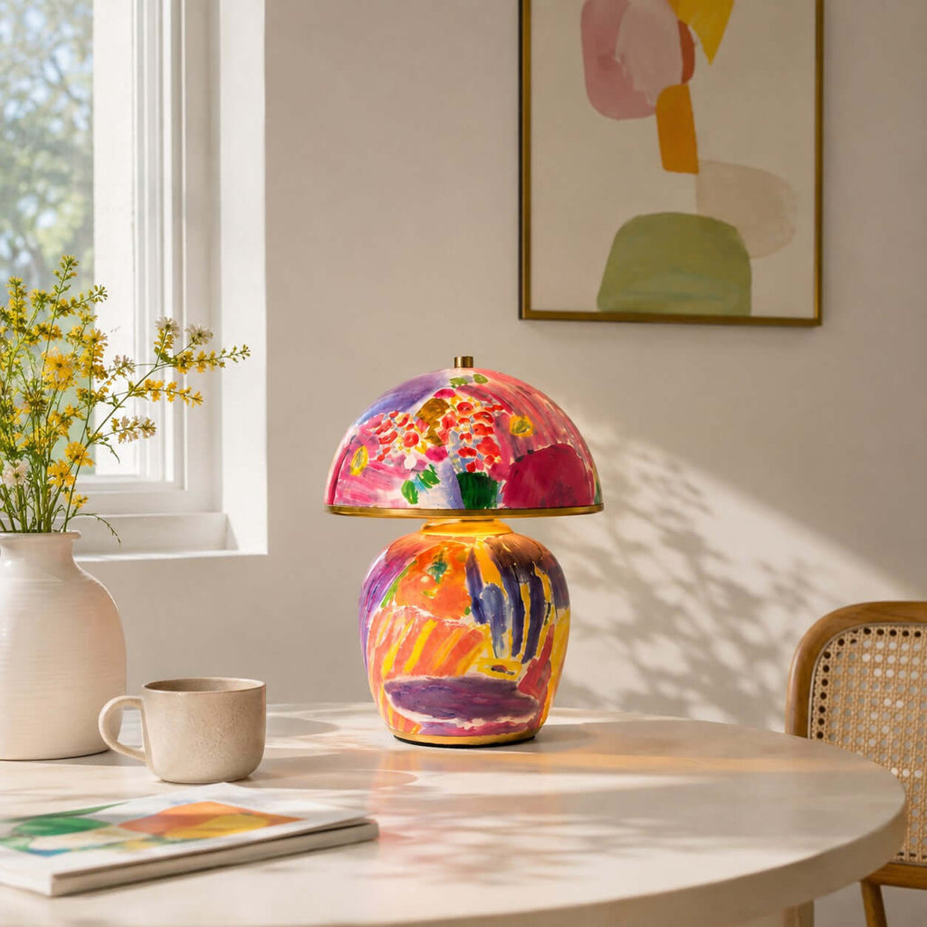 Seen here, a Clérachic table lamp on a round wooden table by a window, with a cream mug, an open magazine and a vase of yellow flowers.