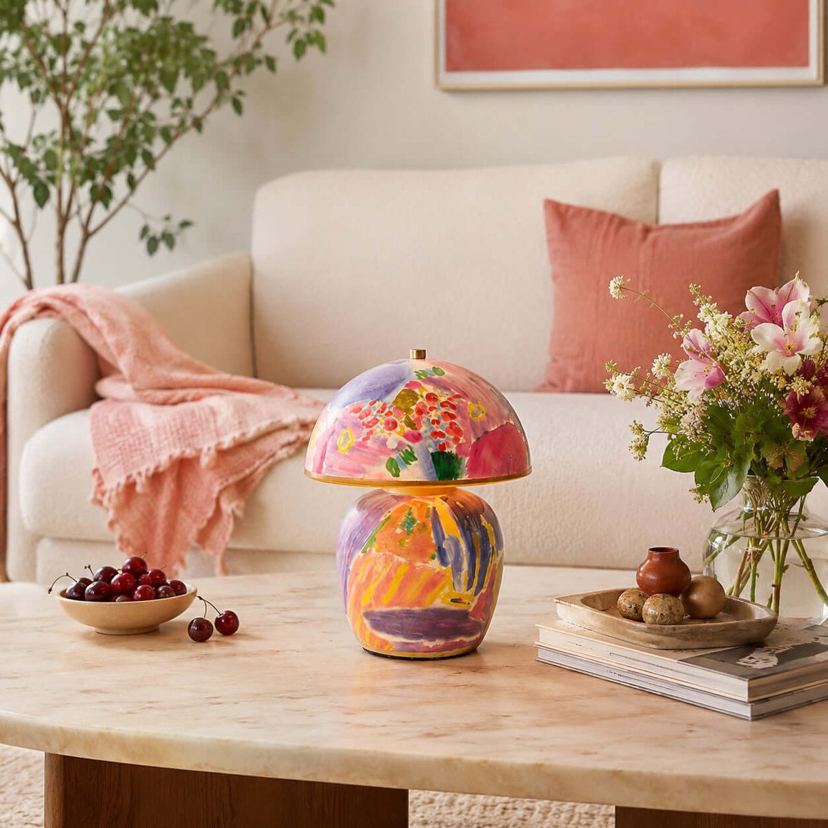 A Clérachic table lamp on a marble coffee table in front of a cream sofa, with a bowl of cherries and a vase of pink flowers.
