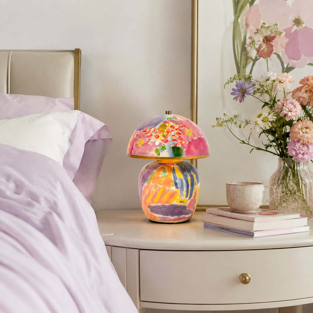 A Clérachic table lamp on a round white bedside table, beside lilac bedding, a mug, a stack of books and a vase of pink flowers.