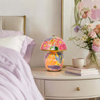 A Clérachic table lamp on a round white bedside table, beside lilac bedding, a mug, a stack of books and a vase of pink flowers.