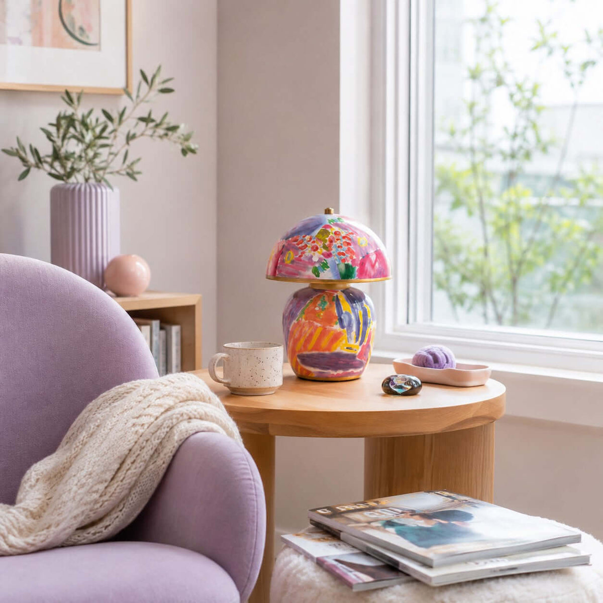 A Clérachic table lamp on a round wooden side table by a sunny window, with a lilac armchair, a mug and a stack of magazines.