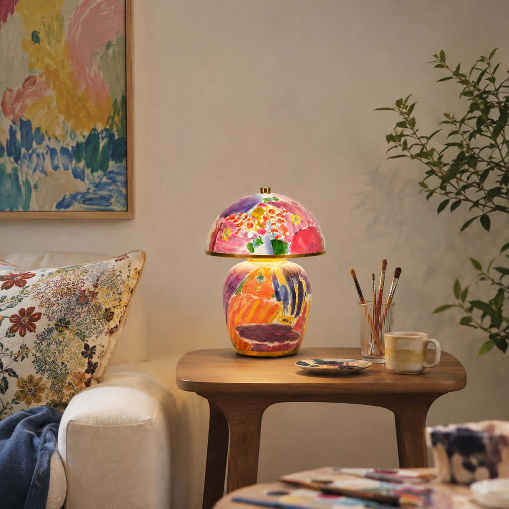 In this photo, a Clérachic table lamp on a wooden side table, with a jar of paintbrushes, a mug and a painted floral canvas behind it.