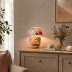A Clérachic table lamp lit on a pale cabinet with brass handles, beside a glass vase of dark pink flowers and a framed print.