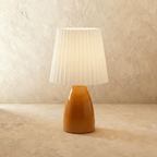The Clérachic ceramic table lamp in a warm orange finish, paired with a pleated white fabric shade, unlit.