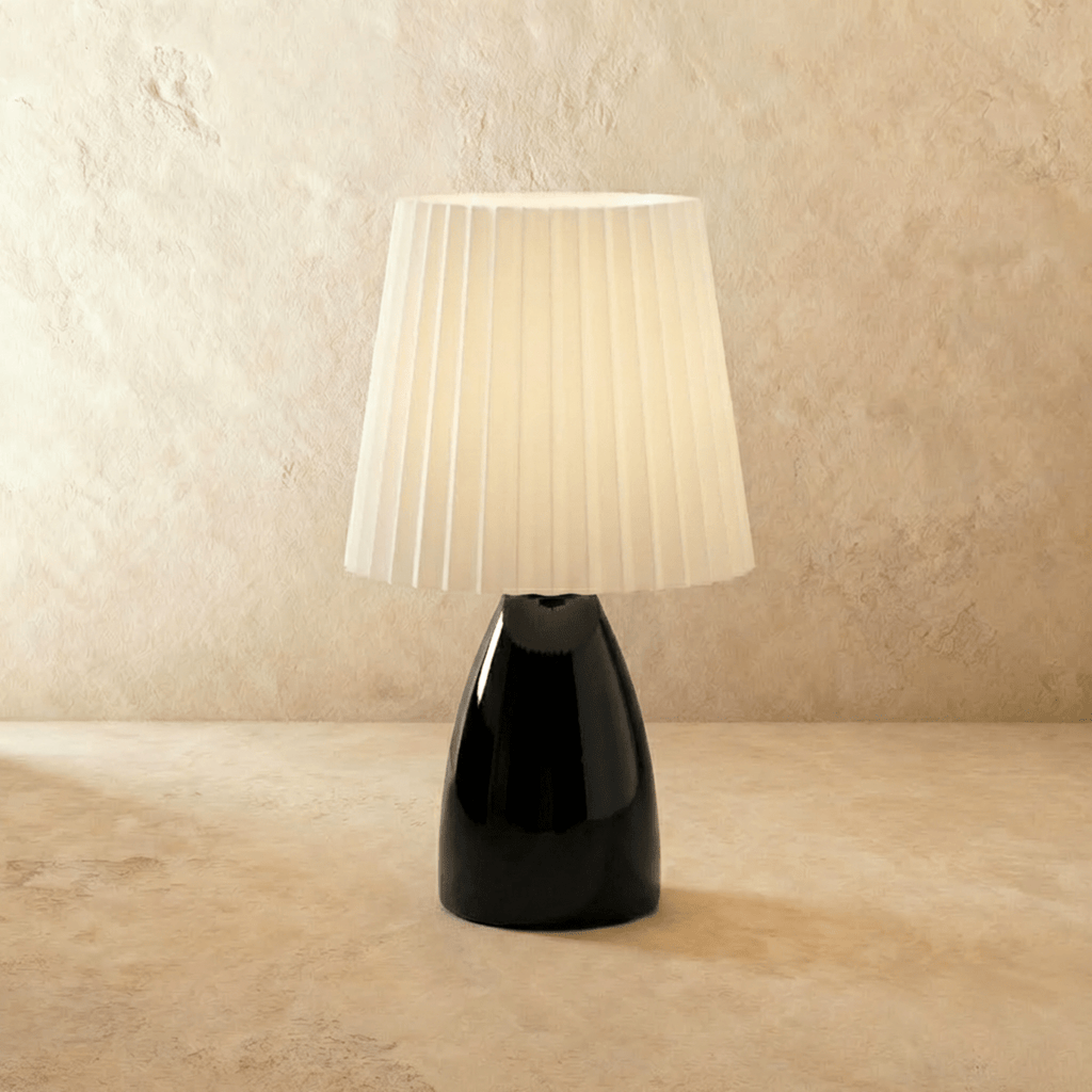The Clérachic ceramic table lamp in glossy black, its pleated white fabric shade glowing warm white.