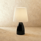 The Clérachic ceramic table lamp in glossy black, its pleated white fabric shade glowing warm white.