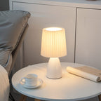 The Clérachic white ceramic table lamp lit on a round side table, next to a teacup, saucer and a rolled magazine.