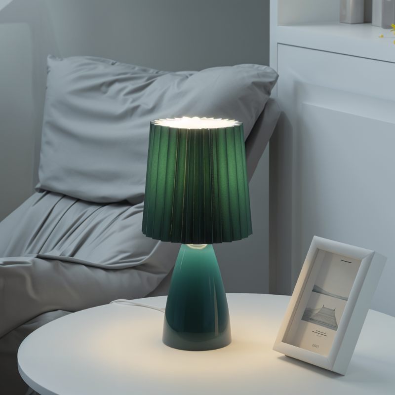 The Clérachic green ceramic table lamp lit on a bedside table, beside a gray upholstered headboard and a framed photo.