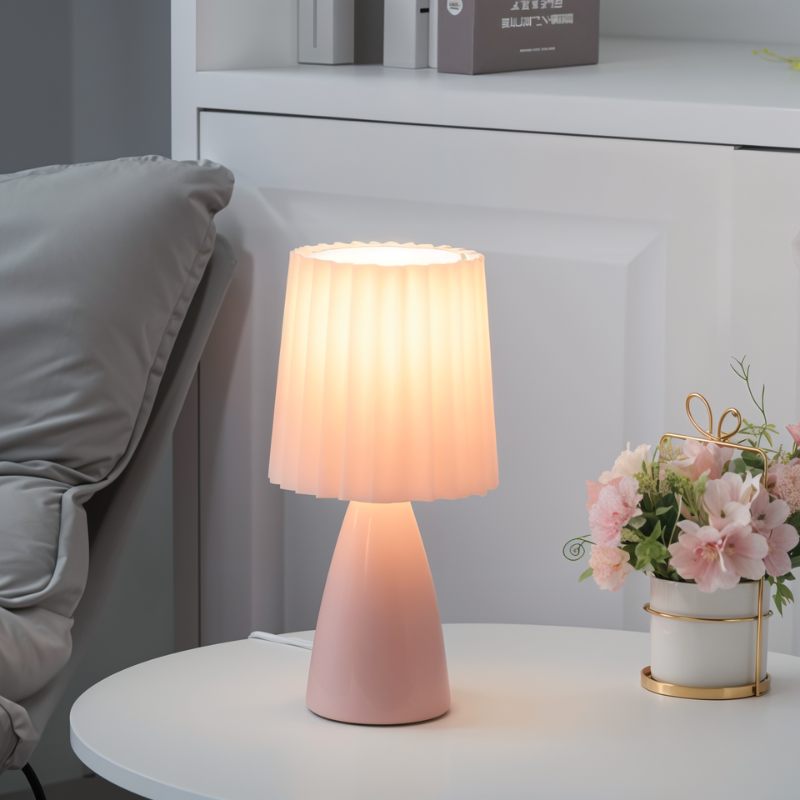 The Clérachic pink ceramic table lamp lit on a white side table, beside a pot of pink flowers and shelved boxes.
