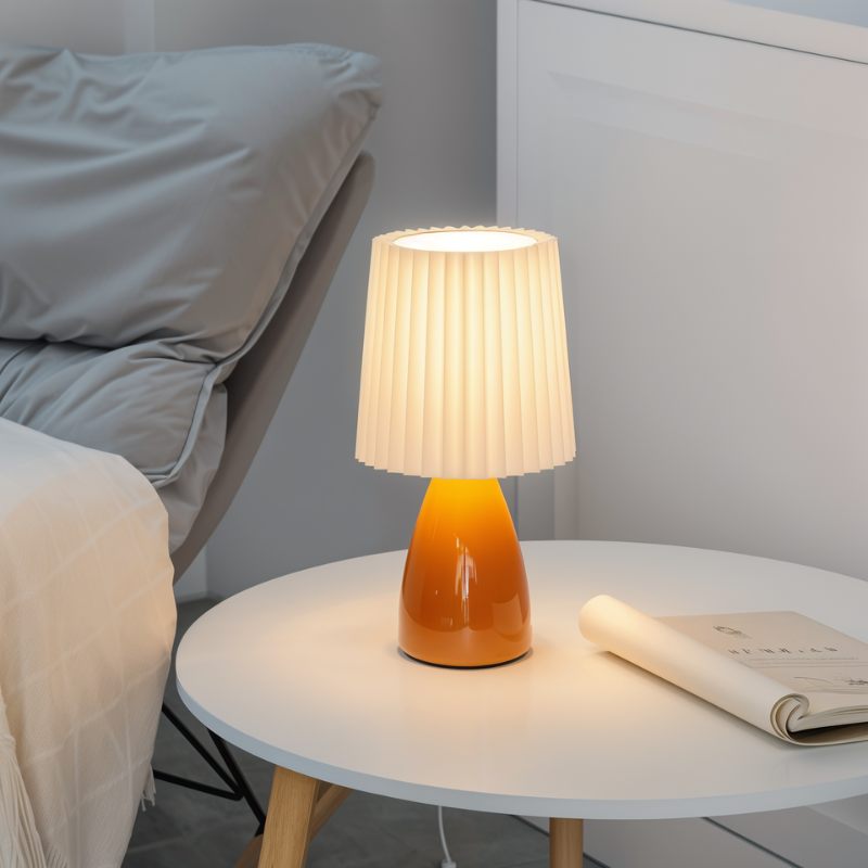 The Clérachic orange ceramic table lamp lit on a wooden-legged side table, beside gray bedding and a rolled magazine.