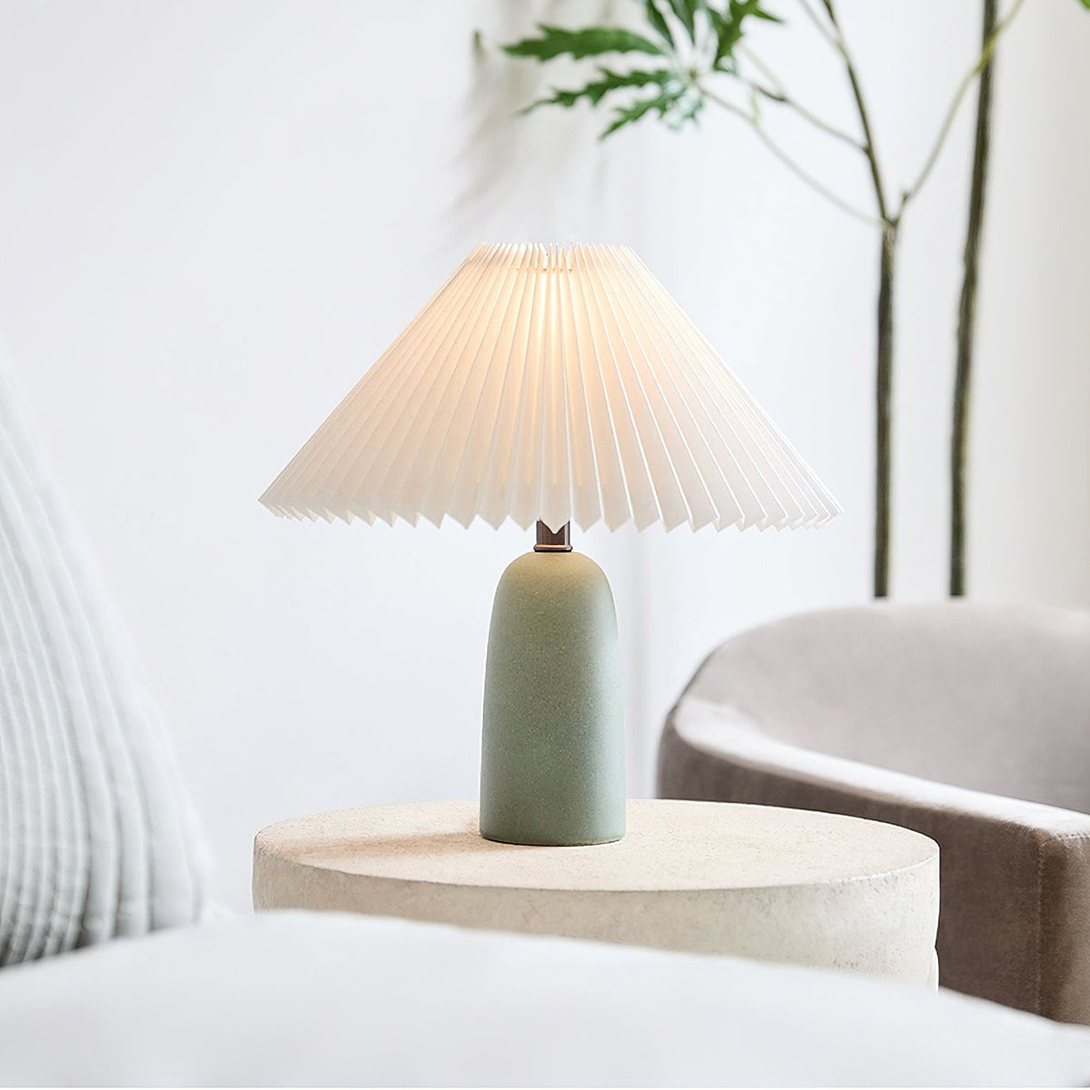 A Clérachic table lamp with a green base and glowing pleated shade sits on a stone-look side table beside a grey armchair and a slim plant.