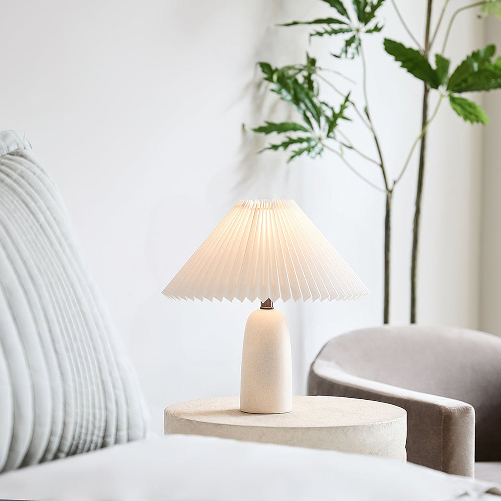 A smaller Clérachic table lamp with a beige base and lit pleated shade on a round stone-look side table, in a bright corner with a plant.