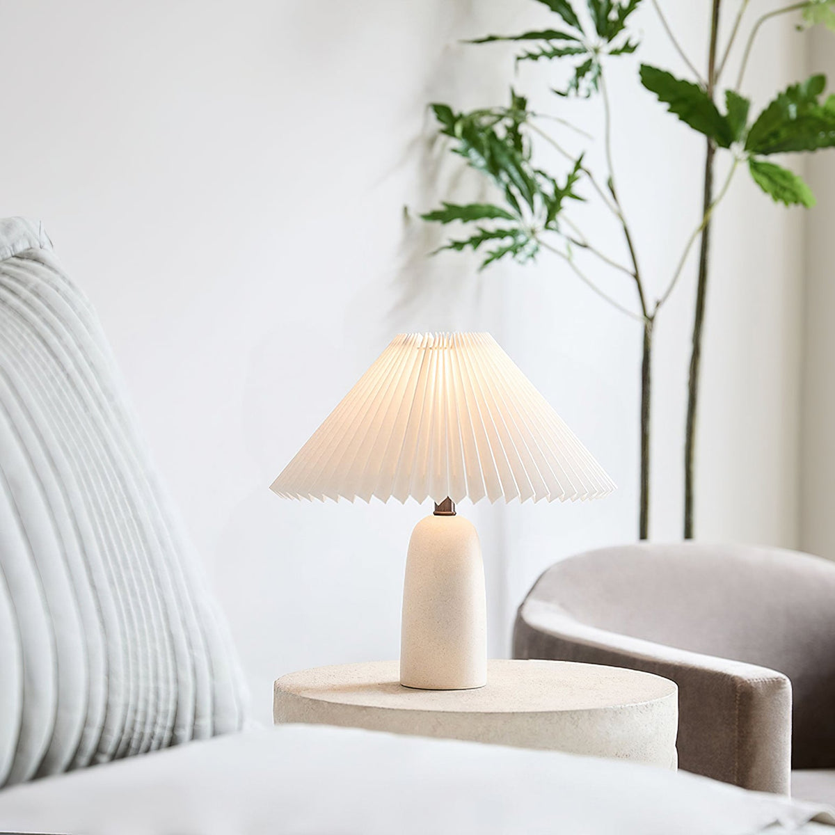 A smaller Clérachic table lamp with a beige base and lit pleated shade on a round stone-look side table, in a bright corner with a plant.
