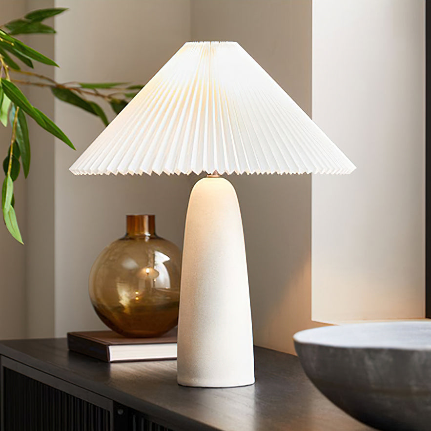 A Clérachic table lamp with a tall beige base and wide pleated shade on a dark sideboard, next to an amber glass vase and a stone bowl.