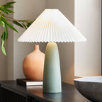 A tall green Clérachic table lamp on a dark sideboard, beside a terracotta vase and a stack of books, under a wide pleated shade.