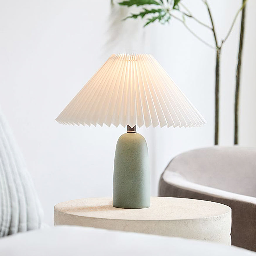 A green Clérachic table lamp with a lit pleated shade on a pale round side table, with a slim plant and a grey armchair behind it.