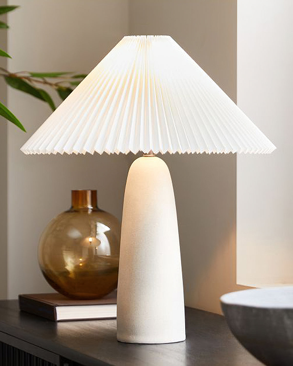 Tall beige base of this Clérachic table lamp on a dark sideboard, with an amber glass vase and the pleated shade glowing above.