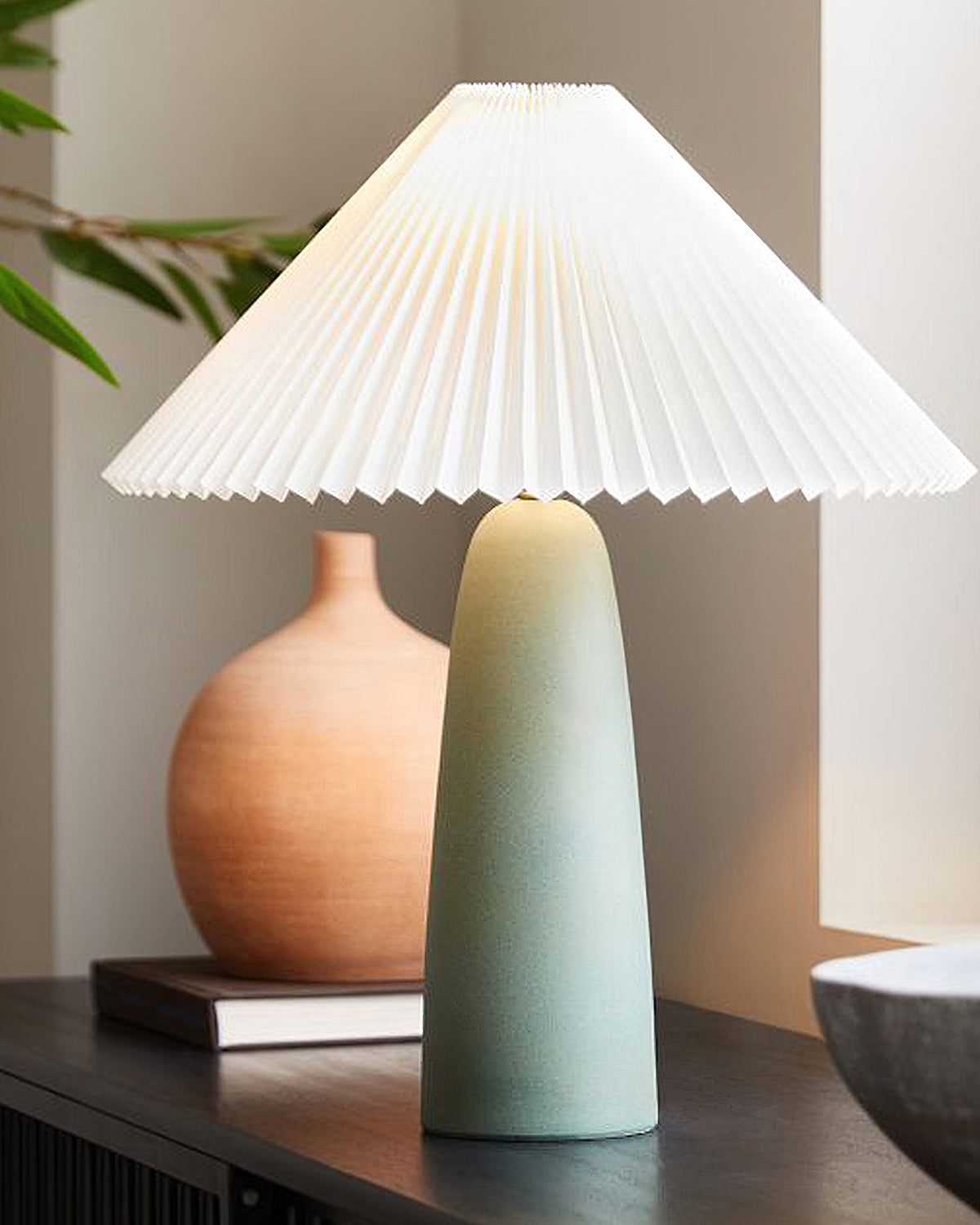 A Clérachic table lamp with a tall green base next to a terracotta vase on a dark sideboard, with leaves behind it.