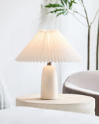 A beige Clérachic table lamp with a lit pleated shade on a stone-look side table, in a bright room with a plant and a grey armchair.