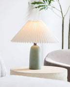 A small green Clérachic table lamp with a glowing pleated shade on a round side table, in a pale room with a tall plant.