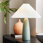A Clérachic table lamp with a tall green base beside a terracotta vase on a dark sideboard, leaves of a plant to the left.