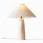 A tall beige Clérachic table lamp lit warm at the top of its pleated shade, on a plain white backdrop.