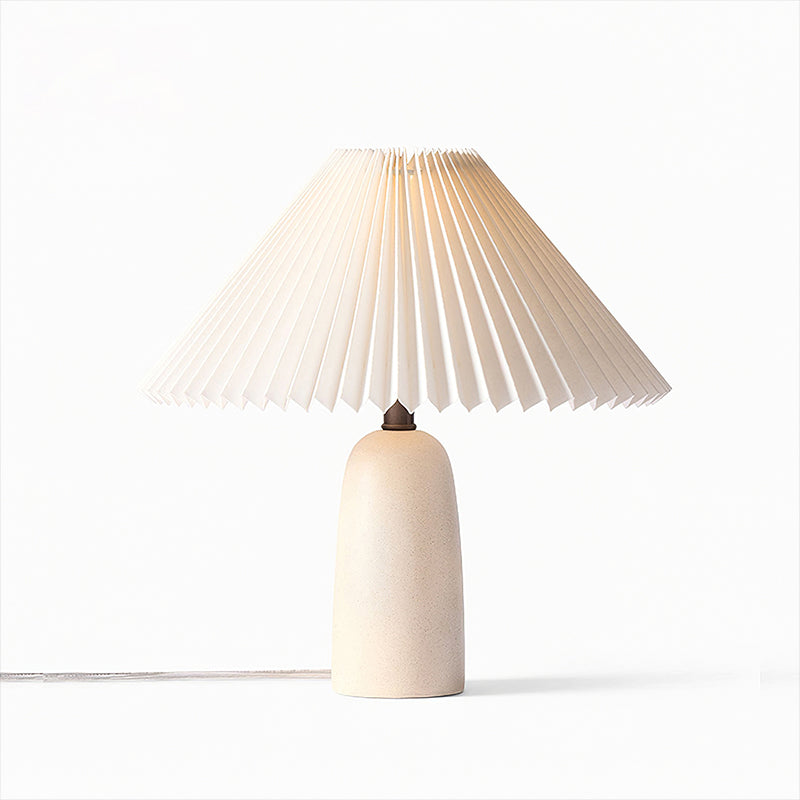 A Clérachic table lamp with a short beige base, lit so the pleated shade glows warm, on a plain white backdrop.
