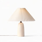 A Clérachic table lamp with a short beige base, lit so the pleated shade glows warm, on a plain white backdrop.