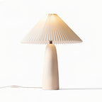 A tall beige Clérachic table lamp lit warm, its pleated shade glowing softly on a white backdrop.
