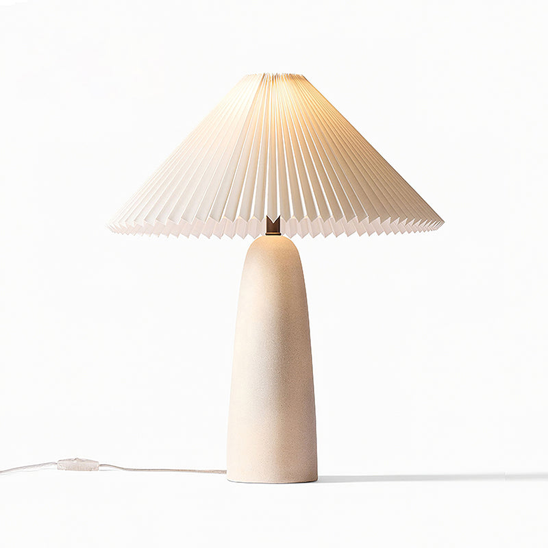A tall beige Clérachic table lamp lit warm, its pleated shade glowing softly on a white backdrop.