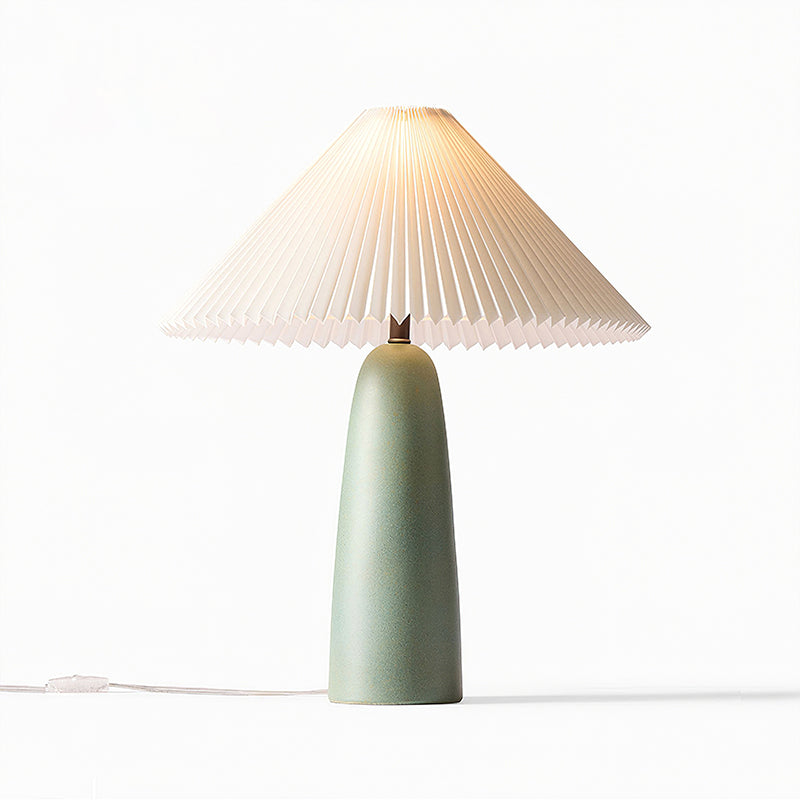 A tall green Clérachic table lamp lit warm, its pleated shade glowing above the matt ceramic base on a white backdrop.
