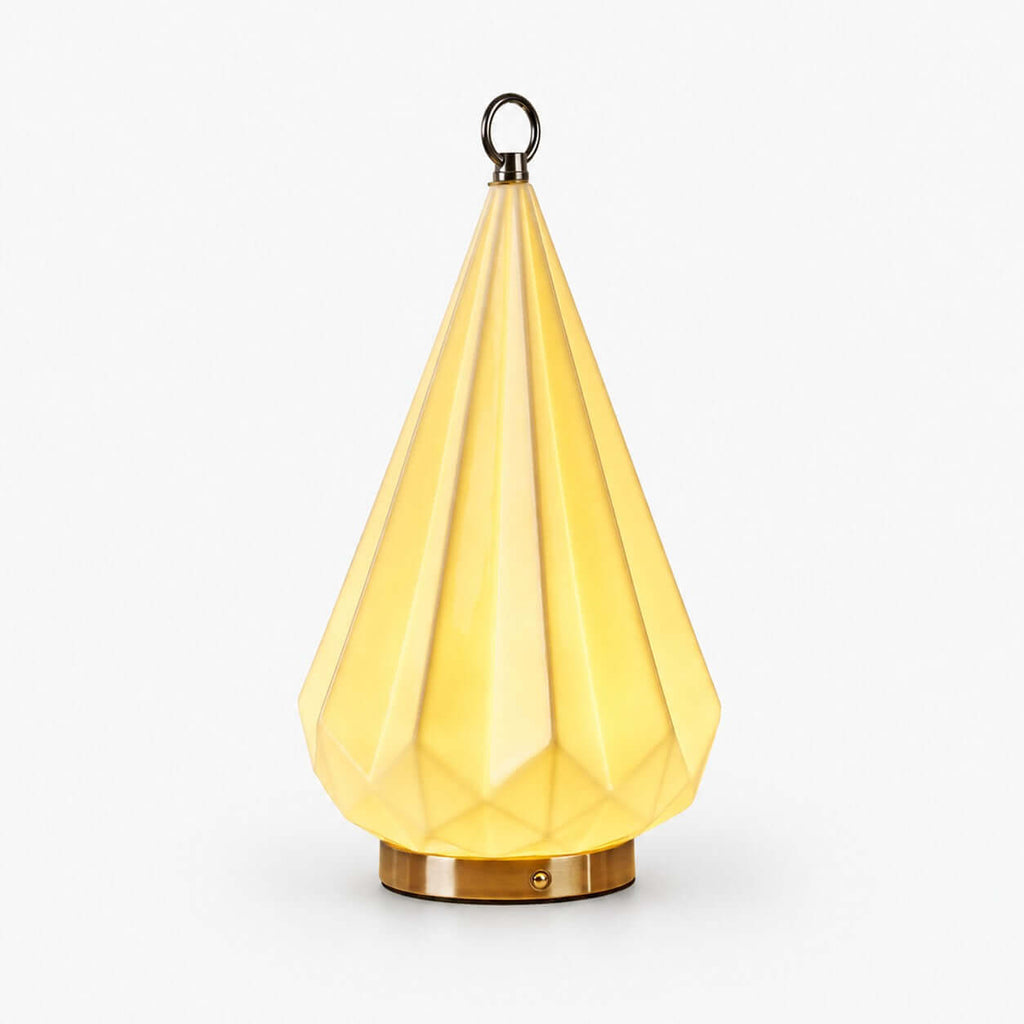 Faceted ceramic table lamp from Clérachic glowing pale yellow, with a brass base and a small metal ring on top, on a white background.