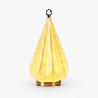 Faceted ceramic table lamp from Clérachic glowing pale yellow, with a brass base and a small metal ring on top, on a white background.