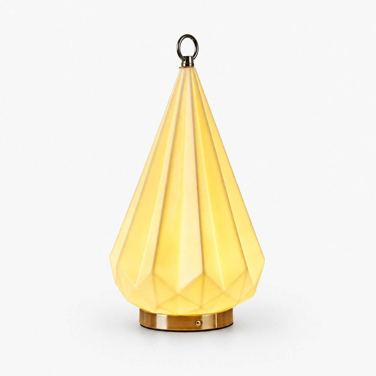 Faceted ceramic table lamp from Clérachic glowing pale yellow, with a brass base and a small metal ring on top, on a white background.