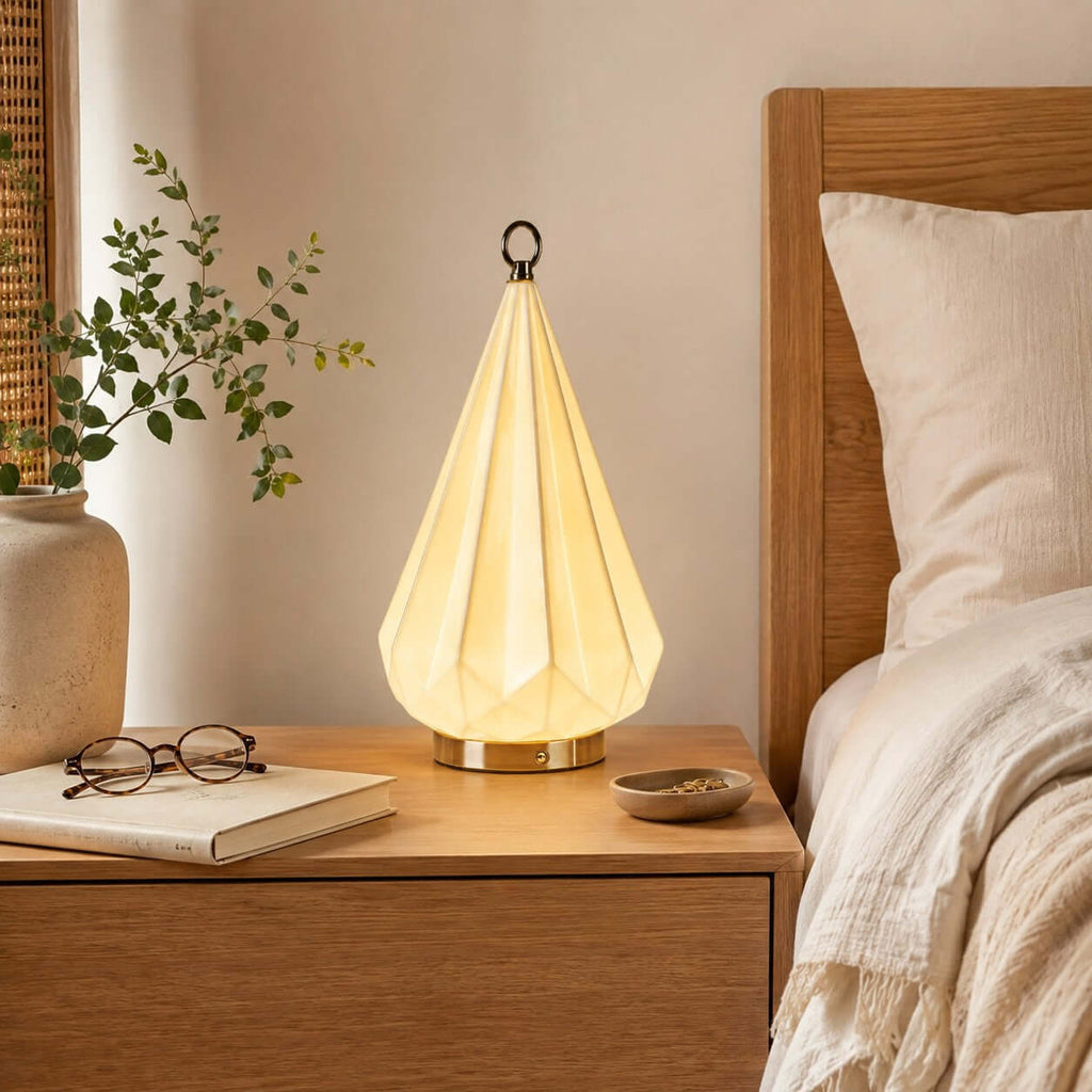 Glowing faceted cone lamp from Clérachic on an oak bedside table, beside a pair of glasses, a book and a cream pillow.