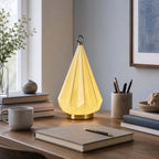 Clérachic faceted table lamp lit on a dark wood desk, next to a stone mug, a notebook with a pen and a pot of pens.