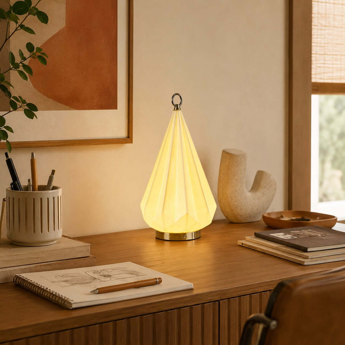 Warm-lit faceted cone lamp from Clérachic on a wooden desk, with a sketchbook, a pencil pot and a sculptural cream vase.